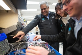 Anantha Chandrakasan smiles and points to a computer chip, alongside two grinning graduate students