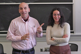 Ken Zolot gestures while standing next to a young woman in front of a blackboard