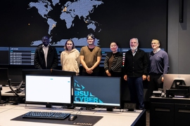 Six people pose standing in front of a wall-sized digital world map. Two computer screens dominate the foreground. One is blank and the other displays “BSU CYBER"