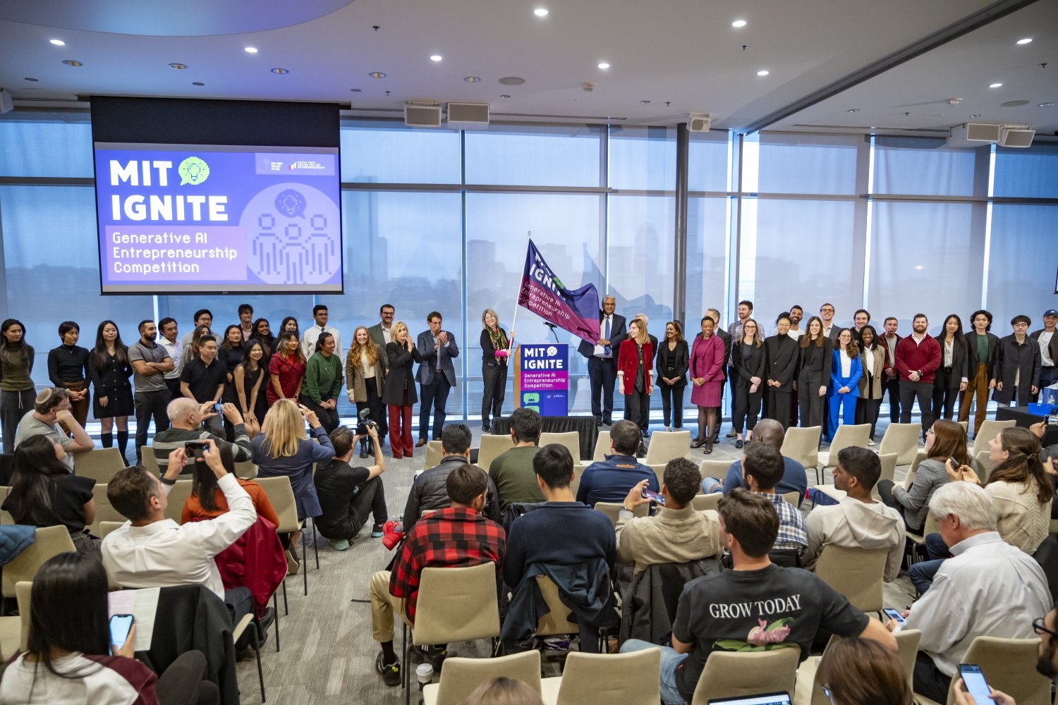 MIT President Sally Kornbluth (left of lectern) and School of Engineering Dean Anantha Chandrakasan (right of lectern) wave the MIT Ignite: Generative AI Entrepreneurship Competition flag alongside other members of Institute leadership and the 12 finalist teams.