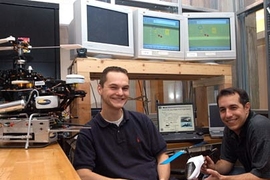 Graduate students Tom Schouwenaars, left, and Mario Valenti sit in front of an operational guidance system they  worked on which is being used  to fly an unmanned aircraft from a manned parent plane to gain surveillance in dangerous situations. They have adapted the same guidance system to the helicopter at left.