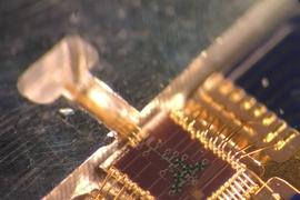 The clock transmitter chip (brown square) wired to a circuit board package. Connected is a metal gas cell (at left), in which a 231.061 GHz signal generated from the chip excites the rotation of carbonyl sulfide molecules. When the molecules reach peak rotation, they form a sharp signal response. That frequency can then be divided down to exactly one second, matching the official time from atomic ...