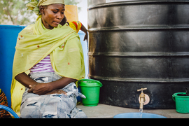 A woman sits next to a large, black metal water container, watching clean water pour out of its spigot