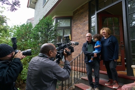 Joshua Angrist, holding his young granddaughter and standing next to his wife, on the steps outside their home, with photographers in the foreground