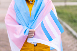 A person is wrapped in the transgender pride flag and is holding a small transgender pride flag.
