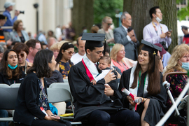 Man in a cap and gown sitting in the audience holds an infant in his lap and kisses him.
