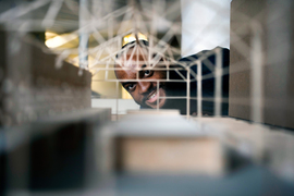 Photo of Justin Brazier smiling as he peeks through an architectural model of a building to the camera