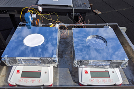 This rooftop photo shows two scales, each holding thick square devices covered in foil. The blue sky is reflected on the foil, and the devices are attached to wires. The left device has a circular indention with white material on top.