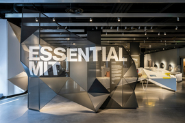 Inside the new MIT Museum, a large grey, angular, see-through sculpture says “Essential MIT” in bold white letters. White tables hold objects in the background.