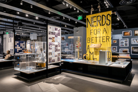 A busy exhibit contains hundreds of objects and photos of MIT history, including a large yellow fabric sign that says “NERDS for a better…” with the last part hidden. An over-sized brass rat ring, a drum connected to a puppet, and a bronze bust of a man with a mustache are visible.