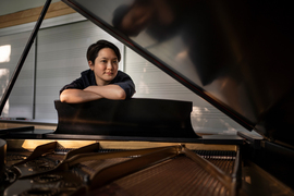 Photo of Jamie Wong, arms crossed on the side of an open grand piano. Piano tines are visible, and the piano's open lid cuts the photo diagonally and frames Wong dynamically.