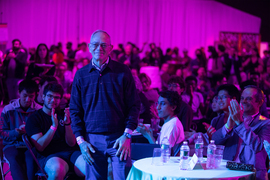 In a large room with at least 100 people in view, MIT President L. Rafael Reif stands as others seated at tables next to him clap. Some in the background are standing. 