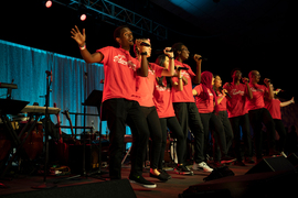 Photo of a diverse group of 9 students, singing on a stage wearing red t-shirts and black pants