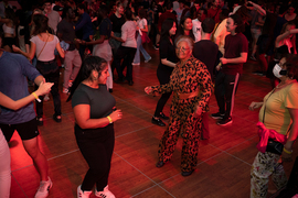 Photo from a large dance party with dozens of people in view, focused on two women, one of whom is wearing a floral 70s-style jumpsuit