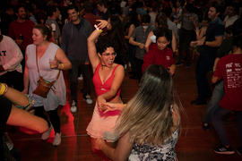 Photo from a large dance party with dozens of people in view, focused on two women holding hands, one of whom has her hands up and a huge smile on her face