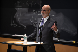 Photo shows solo Tom Wolf speaking with blackboard in background.