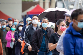 Photo, taken in Hong Kong, shows community members waiting in line, and everyone is wearing a surgical mask. Some have rain jackets or umbrellas.