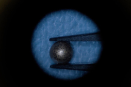 A tiny spherical magnet is held between tweezers in a microscopic photo.