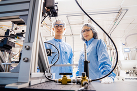 Farnaz Nirouiand and Spencer Zhu in safety glasses and shower caps look down at camera from bright laboratory with a metal machine in the foreground at left