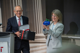 Rafael Reif holds an open box with a red bow, while Sally Kornbluth holds up a blue glass pumpkin. Both are laughing.