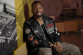 Austen Roberson smiles while seated indoors, and wears a NASA “Black Space” jacket with patches of astronauts Guion Bluford and Mae C. Jemison.