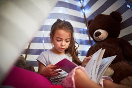 A child reads a book inside a tent with a teddy bear.