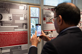 A man takes a photo with his phone while looking at a poster saying, “First South Asian Undergraduates at Tech.”