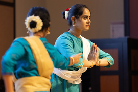 2 dancers with teal garments, gold jewelry and eye makeup, one holding her hands pressed together.