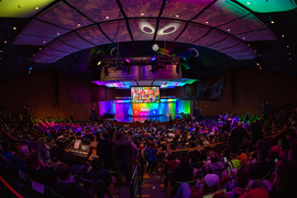 Kresge Auditorium, seats filled with people, lighted with a disco ball and rainbow-colored lights on stage. The stage has a large screen that says “MOVE!” with a picture of the Red Team displayed above it.