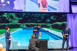 Screen on stage shows swimming pool and prototype SpOtter. One member of the Blue Team demonstrates by immersing their arm with the watch-like device in a water tank while two other Blue Team members stand alongside them.