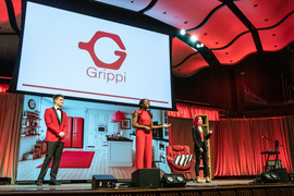 Wearing red and black, three members of the Red Team is on stage with screen behind them showing Grippi logo and a kitchen. The protoype Grippi, a red chair, two black-and-white-striped pillows, and a wooden bookcase with red books are also on stage.