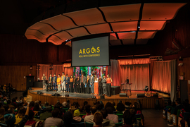 The Orange Team wears orange, grey, navy, and black, and is on stage with screen behind them showing Argos logo and a street with brick houses and streetlights. A wheelchair is also on stage.