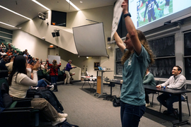 Two students are holding up signs in front of an auditorium of students. Behind them is a screen with projected image of football players. 