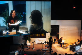 The stage looks like a living room, and two actors read scripts. A camera operator is on stage. A large screen above the stage shows the camera operator’s view of the other two actors.