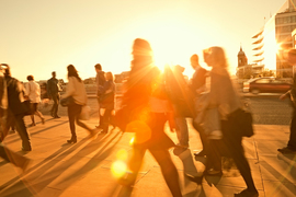 Blurry people walk down a sidewalk with bright orange sun rays creating shadows and lens flare.