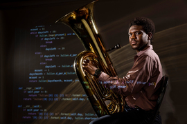 Frederick Ajisafe sits and holds a tuba in a dark room. Coding language is projected onto Ajisafe and the wall.