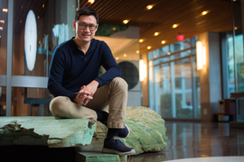 Gene Wei Li smiles and sits on green-tinted rocks inside a building at MIT. Windows and lights are in the background.
