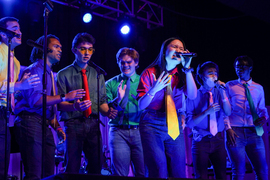 On stage, Ji steps forward and sings dramatically. About seven Logarythms members stand in background. Everyone wears colorful ties and blue jeans.