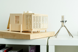 Two architectural models. On left, a wooden boxy folding home with patterned windows, and on right a metal tripod-like support device.