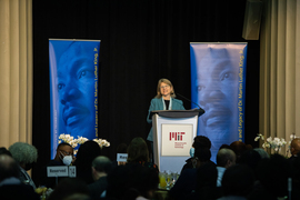 Sally Kornbluth speaks at the podium, with two blue banners featuring the face of Martin Luther King on either side.