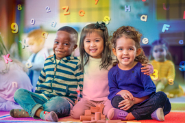 3 smiling preschoolers sit down for a group photo inside a classroom. Letters and numbers swirl around the colorful air.