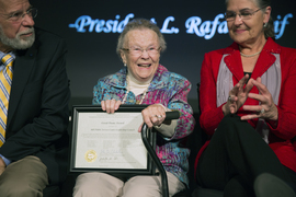 Priscilla smiles while sitting on stage between two people. She holds a framed award that says “Great Dome Award.”