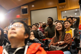 Seated individuals in auditorium react with various expressions, including jubilant and serious, as they watch the competition.