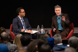 Dozier, left, and Tomasi in conversation in front of an audience while seated in red chairs.