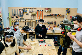 In a busy wood-working lab, 7 teachers and students wear safety masks and goggles as they work with wood. Lots of tools and equipment can be seen on the walls.