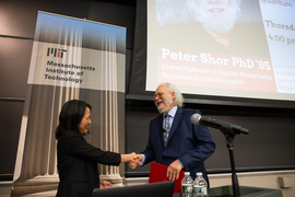 Lily Tsai shakes hands with Peter Shor on the presentation stage. Shor is holding a red folder, and both are smiling.