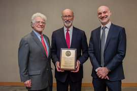 The three men wear suits and pose for a photograph. Wolf holds his award plaque that has the MIT seal on top and small text below.
