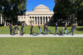 14 members of the research team pose the word “WORMS” (four as the letter “W”, three as the letter “O”, two as the letter “R”, three as the letter “M”, and two as the letter “S”) in grassy area in front of MIT’s dome.