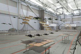 Combined video stills show the pathways of four flying drones in a large auditorium. The four drones avoid two flying obstacles, which are drones with a camouflage box around then.