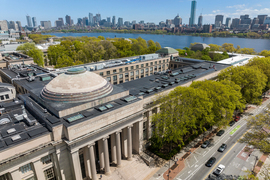Aerial view of the MIT campus shows the roofs of several buildings, including the Great Dome, with a glimpse of Mass Ave, the Charles River, and the Boston skyline in the background.